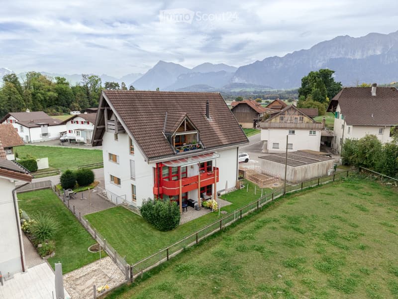 A two-story house with a red balcony, surrounded by a well-maintained lawn and garden, with neighboring houses and a scenic mountain backdrop.