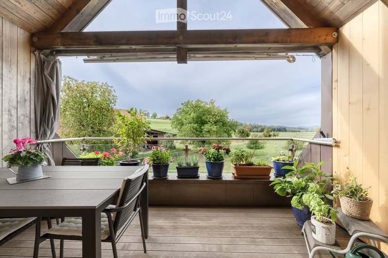 Wooden balcony with a table, chairs, and several potted plants, overlooking a scenic countryside view.
