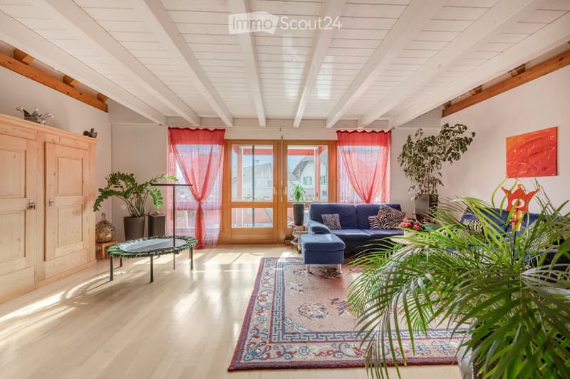 Bright living room with wooden floors, high ceilings, large windows with red curtains, blue sofa, ottoman, side table, and decorative plants.