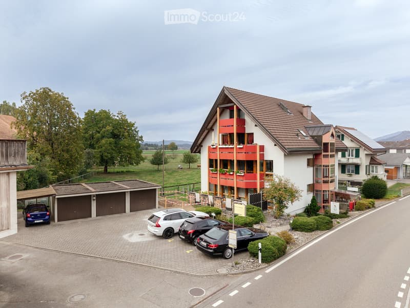 Two-story house with red-tiled roof, white walls, and red balconies. Garage with multiple cars parked in front, surrounded by greenery and open fields.