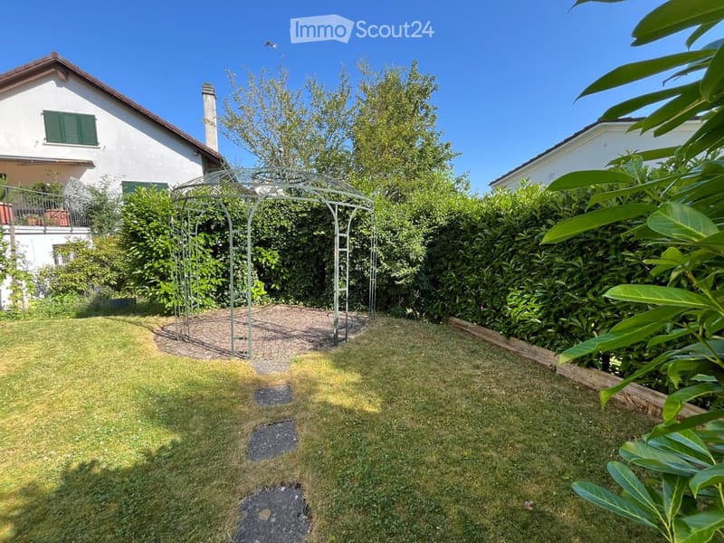 Well-maintained garden with a lawn, a metal archway, and a house with green shutters and a balcony in the background.