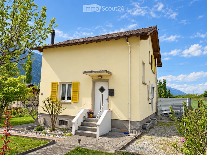 Single-story yellow house with white trim, raised ground floor, small porch, and garden area with a bench and plants.