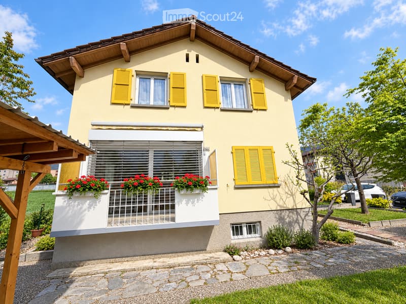 Two-story house with yellow shutters, white window frames, and a balcony with red flowers. The house has a stone base and a wooden roof.