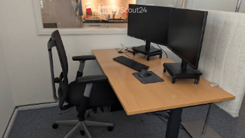 Office space with a wooden desk, ergonomic chair, dual monitors, keyboard, and mouse. The room has a grey carpeted floor and white walls.