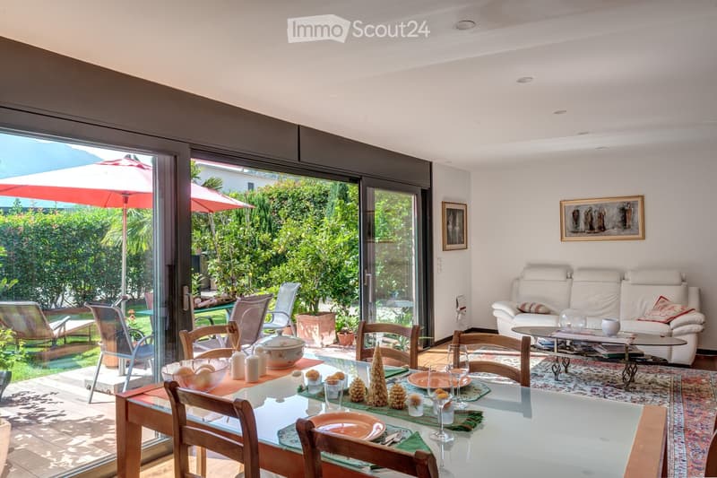 Bright living and dining area with large glass doors opening to a garden, featuring a white sofa, coffee table, dining table with chairs, and decorative items.