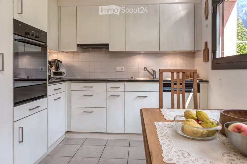 Modern kitchen with white cabinets, stainless steel appliances, a wooden dining table with a lace tablecloth, and a bowl of fruit on the table.