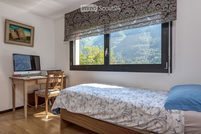Cozy bedroom with a single bed, wooden desk, chair, computer, and a scenic mountain view through a large window.