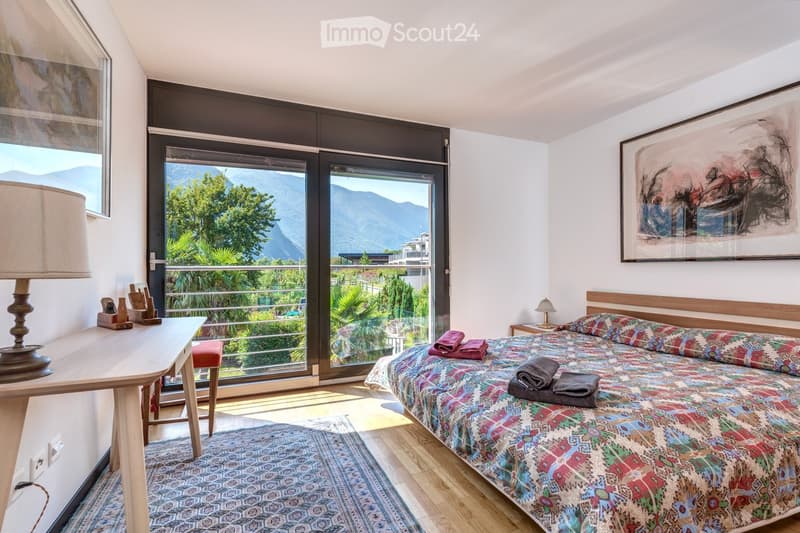 Bright bedroom with large windows offering a scenic mountain view, featuring a colorful bedspread, a wooden nightstand with a lamp, a small table with a lamp, and a framed artwork on the wall.