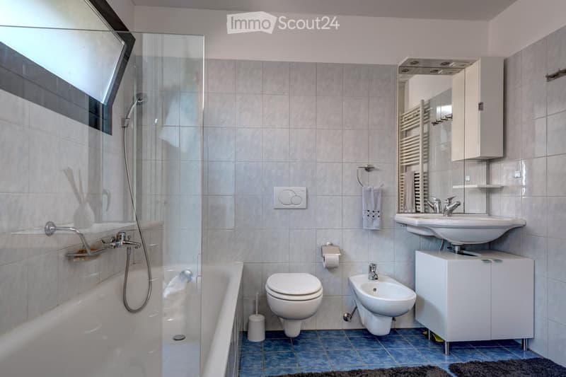 Modern bathroom with a bathtub, toilet, bidet, sink, mirror, and cabinet. Features a glass-enclosed shower area, tiled walls, and a towel rack with a towel.