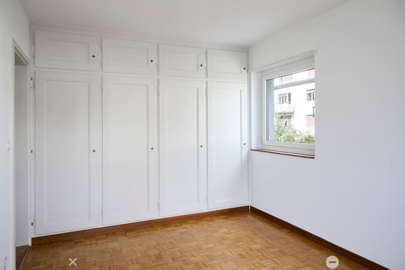 Empty room with white walls, wooden herringbone flooring, built-in white cabinetry, and a window with a view of a neighboring building.