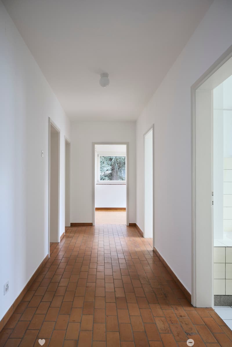 A long, narrow hallway with white walls, wooden tiled flooring, and several doors leading to other rooms. The ceiling is white with a single light fixture.