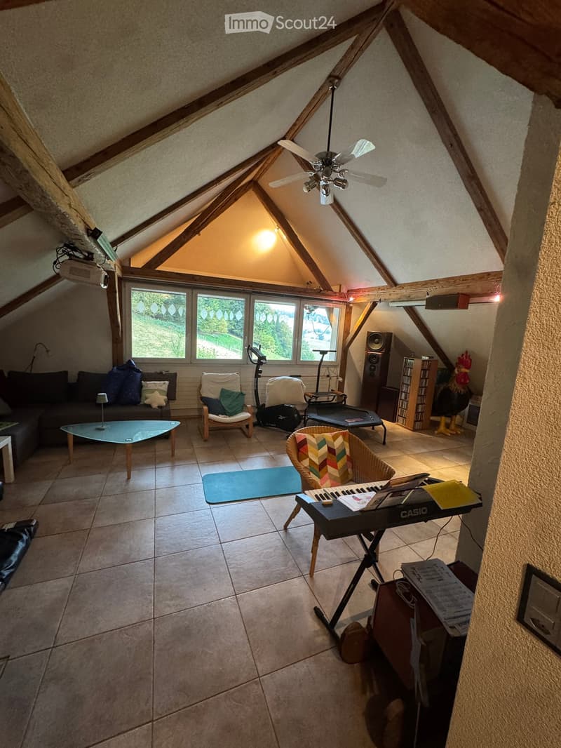 Loft-style living room with exposed wooden beams, large windows, sofa, coffee table, chairs, keyboard, speaker, and decorative items.