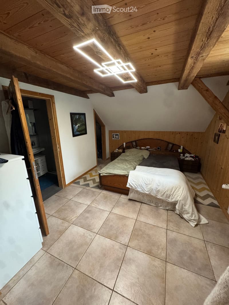 Cozy attic bedroom with wooden beams, tiled floor, bed with white and green bedding, nightstand, framed picture, and an open closet area.