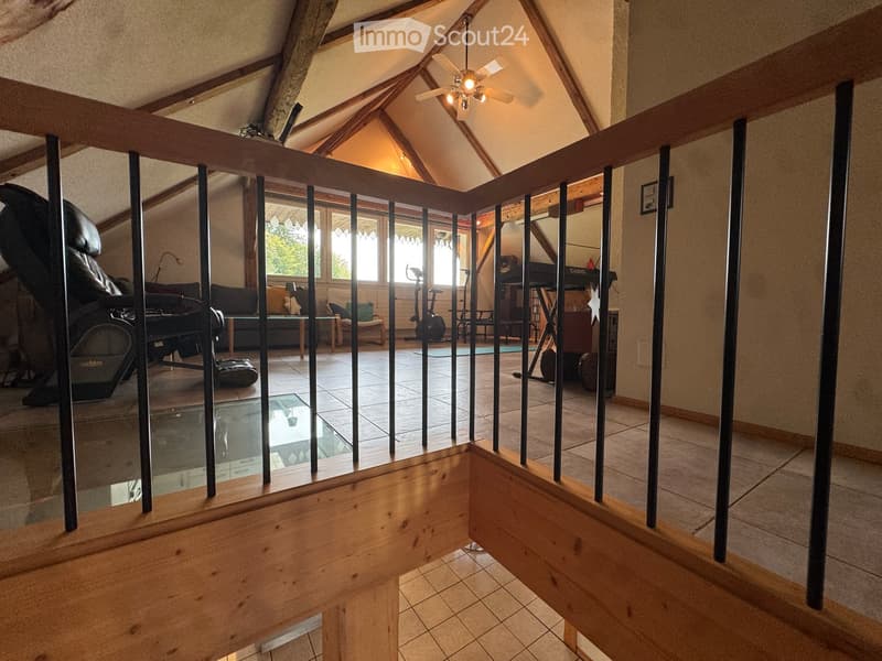 A spacious, well-lit living area with wooden beams, a wooden railing, and large windows. The room features a sofa, chairs, a table, a lamp, a piano, and an exercise bike.