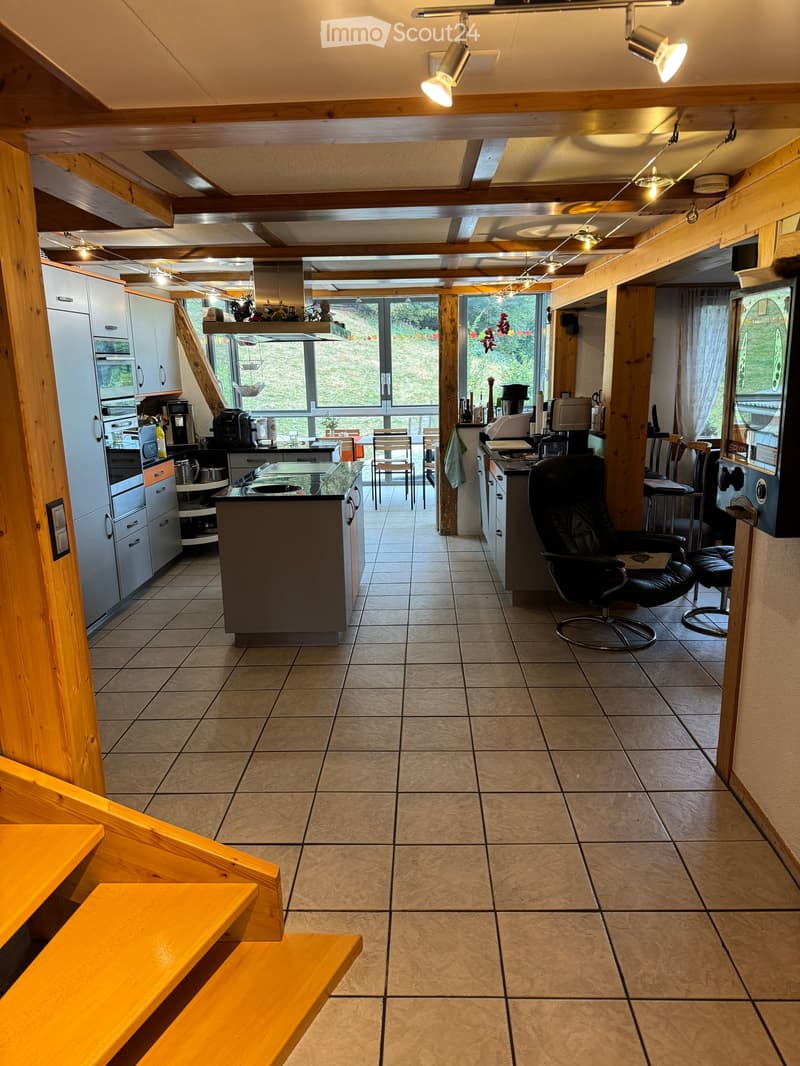 Open-plan kitchen and living area with exposed wooden beams, tiled floor, and large windows overlooking a green area. Kitchen features a central island, cabinets, and appliances, while the living area includes seating and a TV.