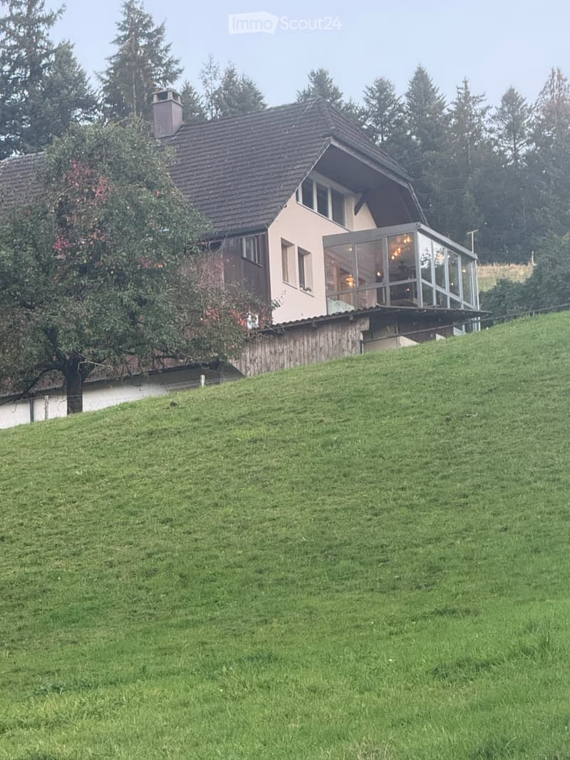 Two-story house with a sloped roof, beige exterior, and a glass-enclosed balcony on the upper level, surrounded by greenery and trees.