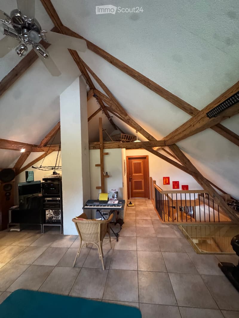 Loft-style room with exposed wooden beams, tiled floor, wicker chair, keyboard on table, stereo system, and glass table near staircase.