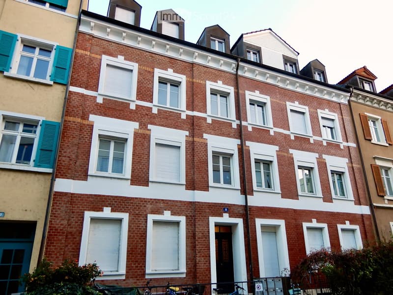 A multi-story brick building with white trim around windows and doors, featuring multiple windows with shutters and blinds, and a small balcony area with bicycles parked below.