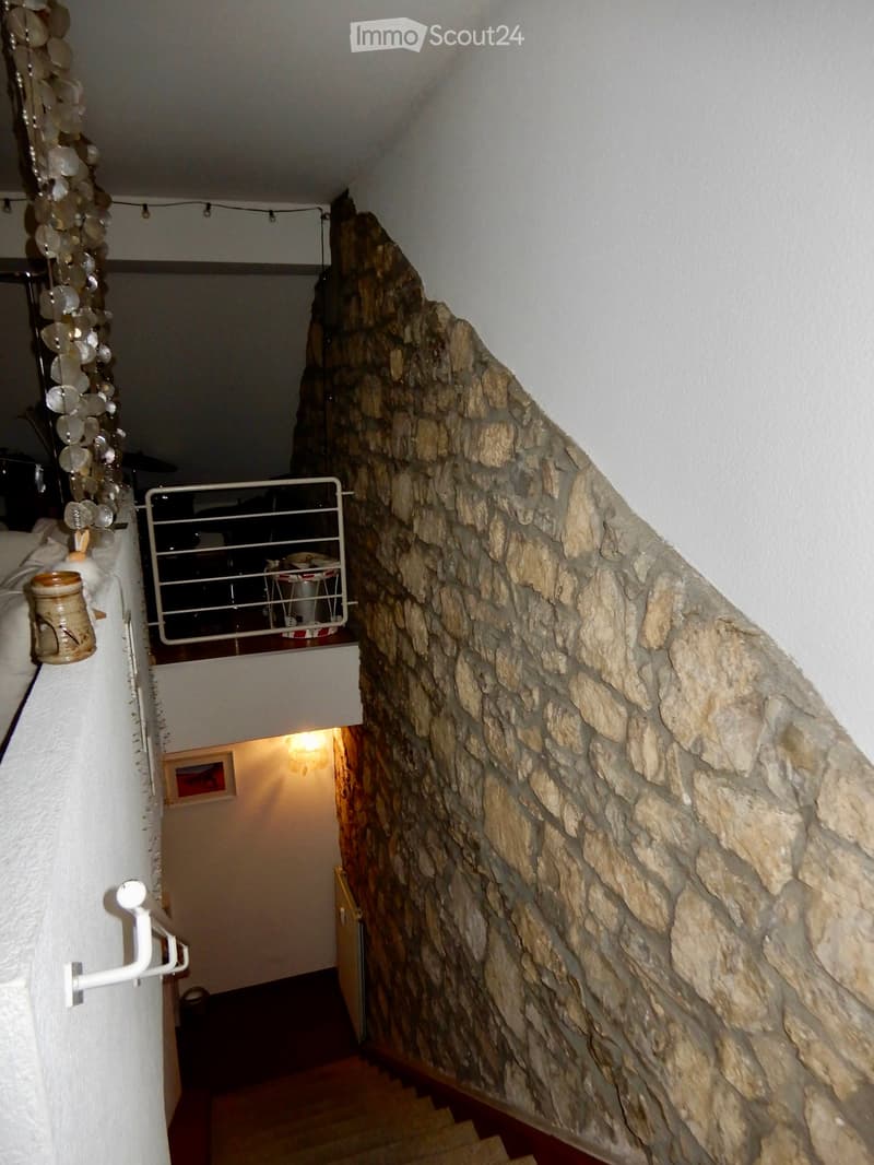 A narrow, steep staircase with wooden steps and a stone wall on one side, leading to a lit room below.