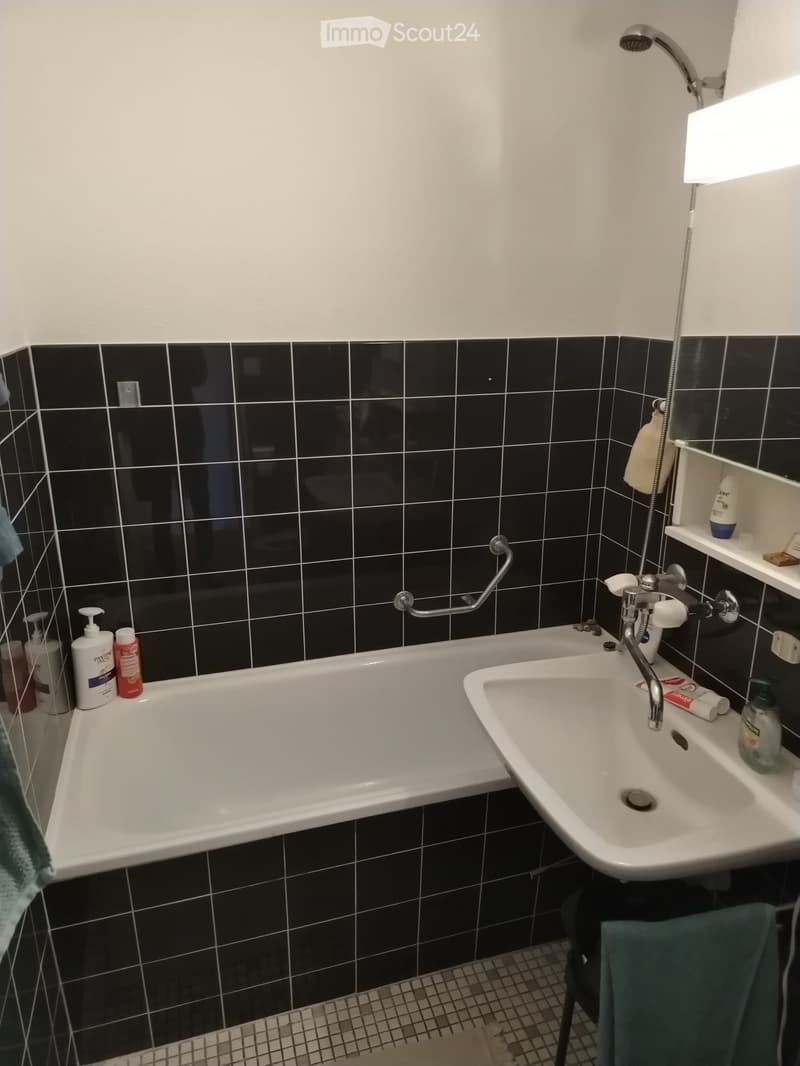 Bathroom with black tiled walls, a white bathtub, a white sink, a showerhead, a handrail, and various toiletries.