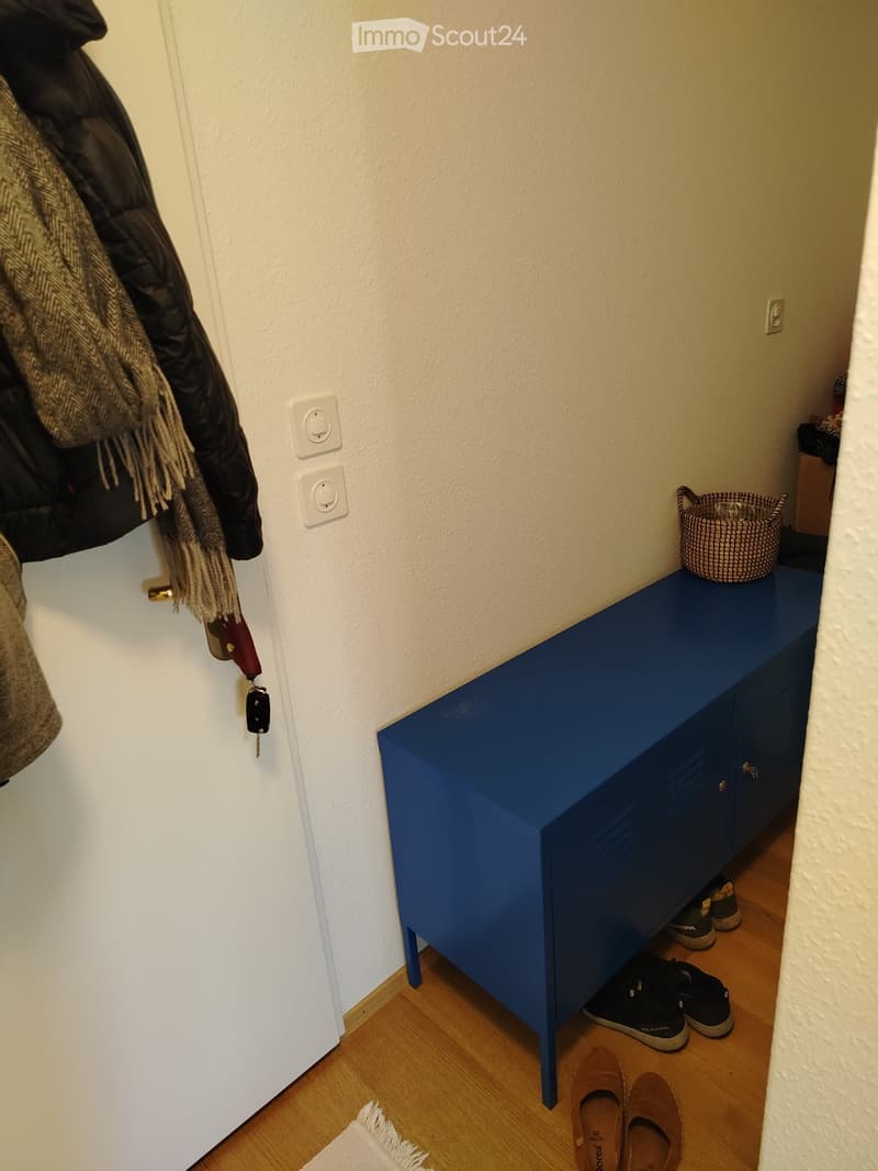 A narrow hallway with a blue bench, a coat rack with jackets and scarves, and a small woven basket on top of the bench. Wooden flooring and white walls are visible.