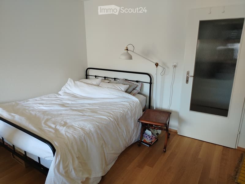 Bedroom with a wooden floor, a bed with white bedding, a small wooden stool, and a wall-mounted lamp.
