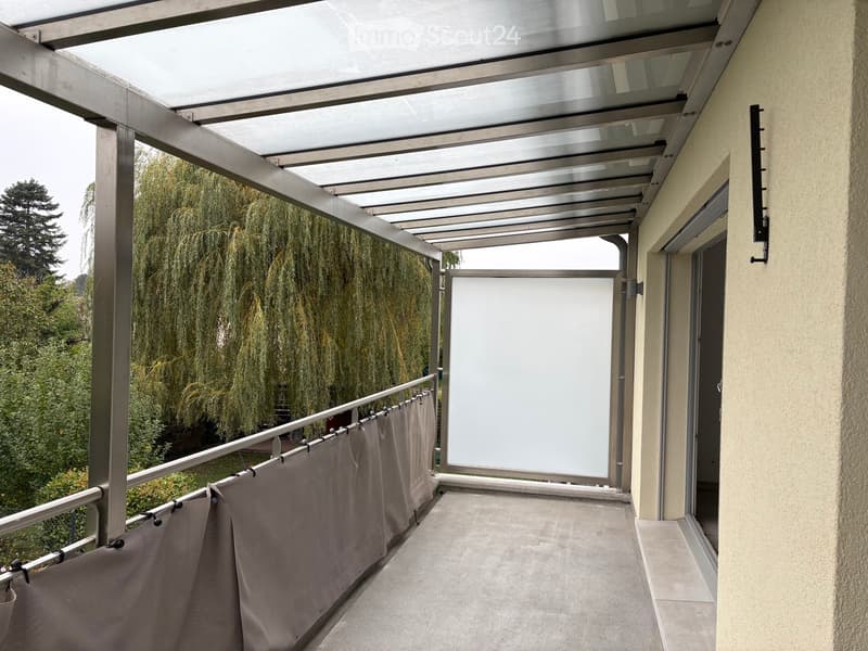 Balcony with a modern metal roof structure, glass panels, and a sliding door leading inside. The balcony has a tiled floor and a metal railing with curtains.