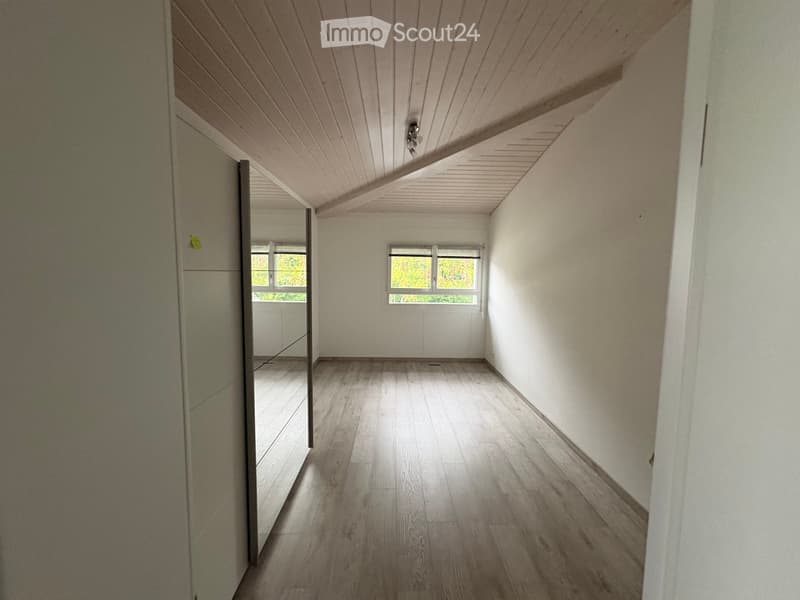 Empty hallway with wooden flooring, white walls, and a slanted wooden ceiling. Two windows provide natural light, and a door leads to another room.