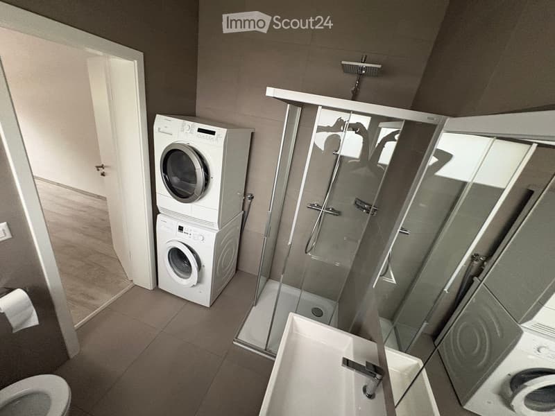 Modern bathroom with a toilet, sink, shower, washing machine, and mirror. The shower has a glass enclosure, and the washing machine is stacked with a dryer.