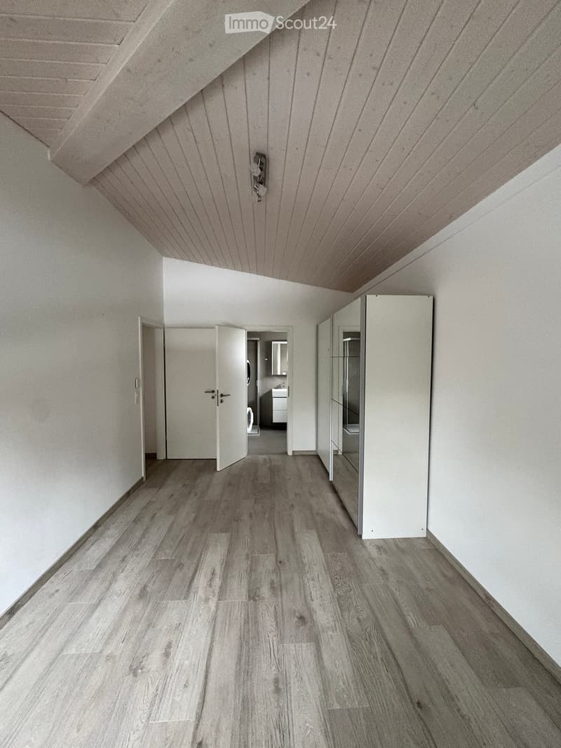 Empty hallway with wooden flooring, white walls, and a slanted ceiling. Doors lead to other rooms, including a bathroom visible at the end.