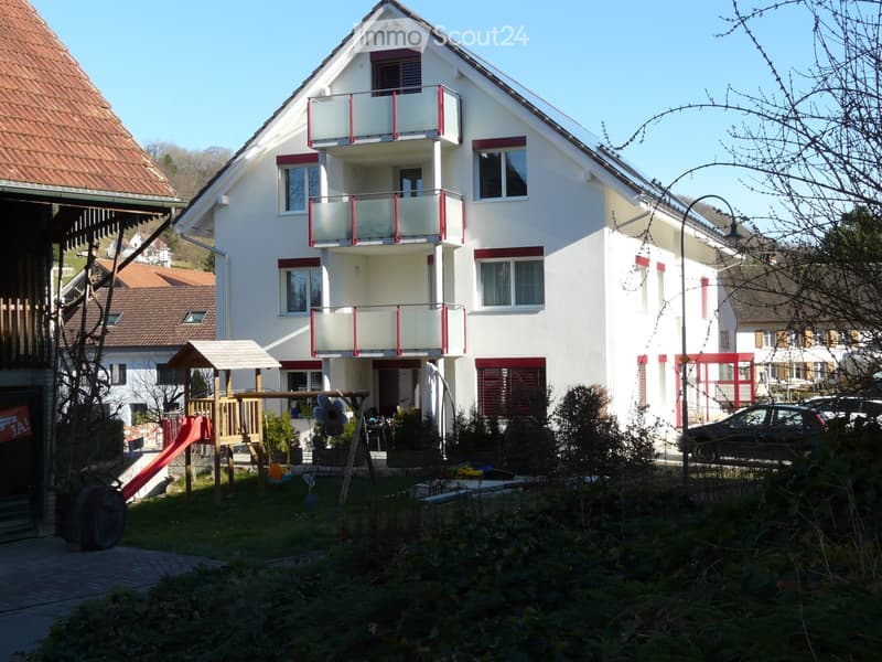 Two-story white house with red trim, multiple windows, balconies, and a small garden area with a swing set and slide.