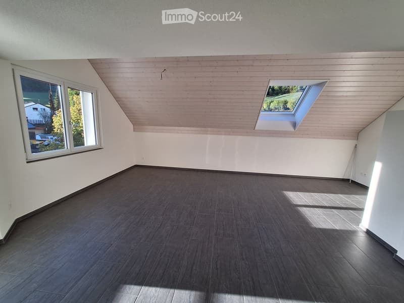 Empty room with wooden flooring, white walls, and a slanted wooden ceiling. Features large windows and a skylight, allowing natural light to enter.