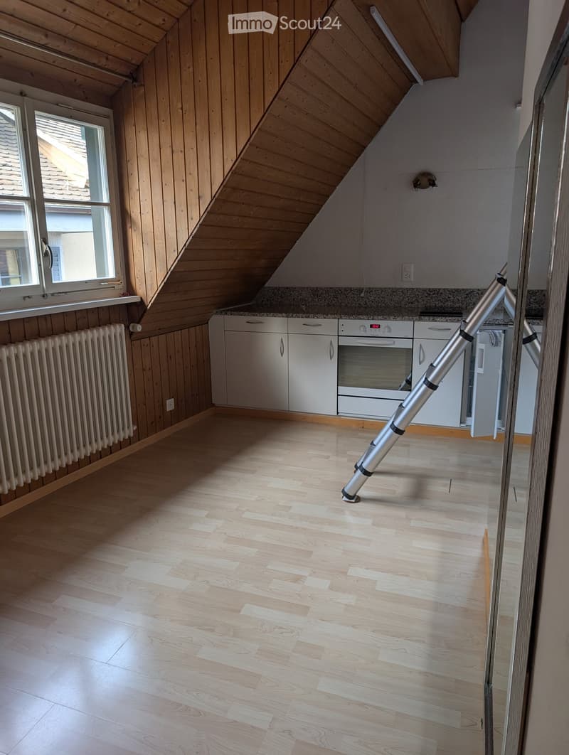 Empty attic room with wooden walls, slanted wooden ceiling, light wooden flooring, and a window. White cabinets and an oven are visible but no furniture is present.