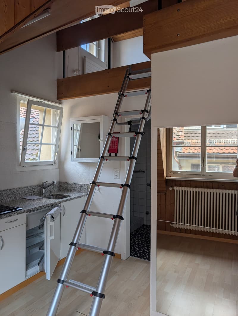 A modern kitchen with a sink, faucet, cabinets, and a mirror, with a ladder leading to an attic space visible through an opening in the ceiling.