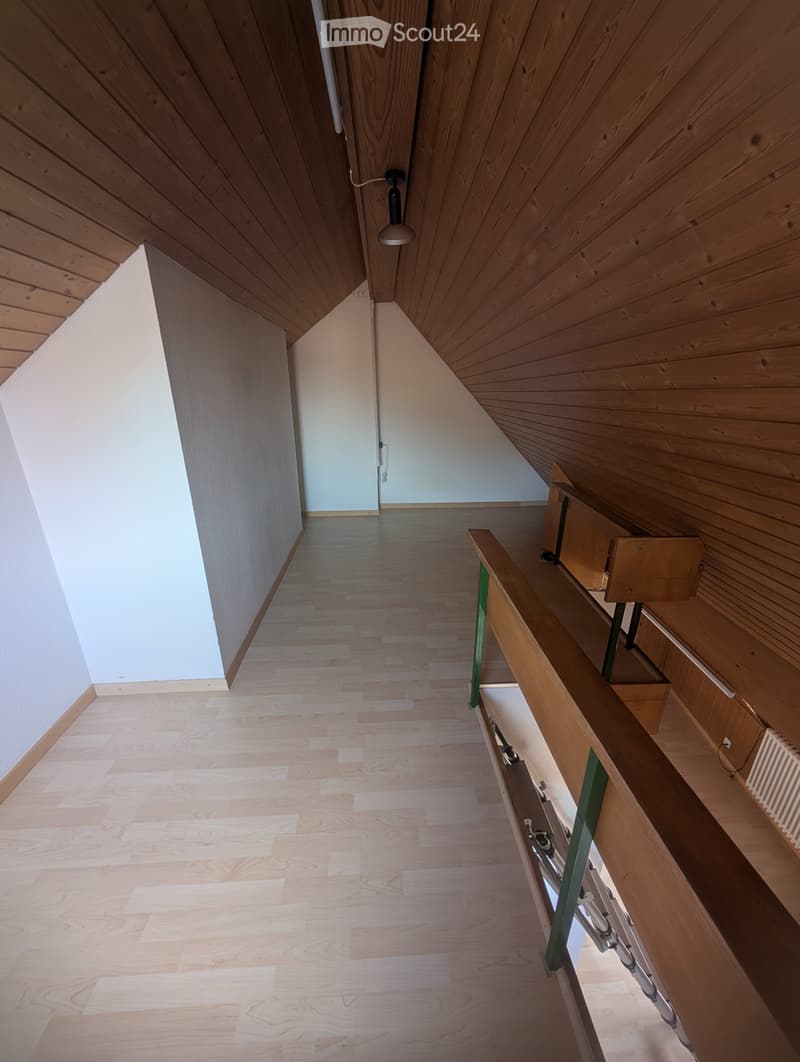 A modern hallway with wooden flooring, slanted wooden ceiling, and a wooden railing on one side. The walls are painted in a light color, and there is a door at the end of the hallway.