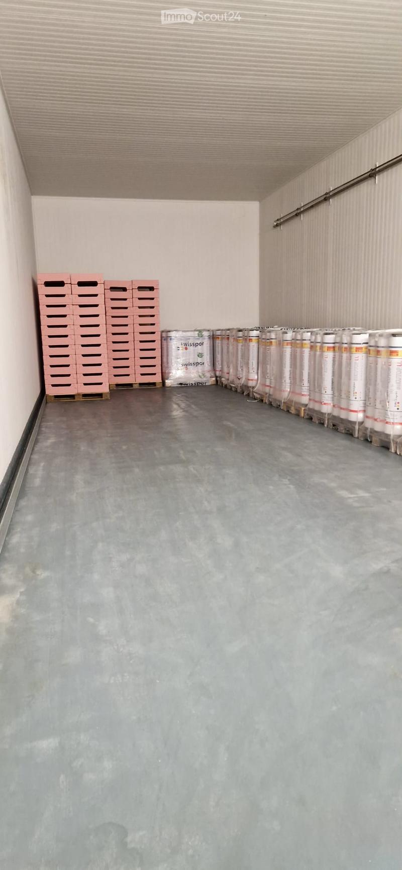 Empty storage room with concrete floor, white walls, and stacked pink crates and wrapped items along one wall.