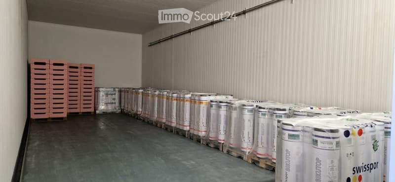 Empty warehouse space with concrete floor, white corrugated metal walls, and stacks of rolled materials and wooden pallets.