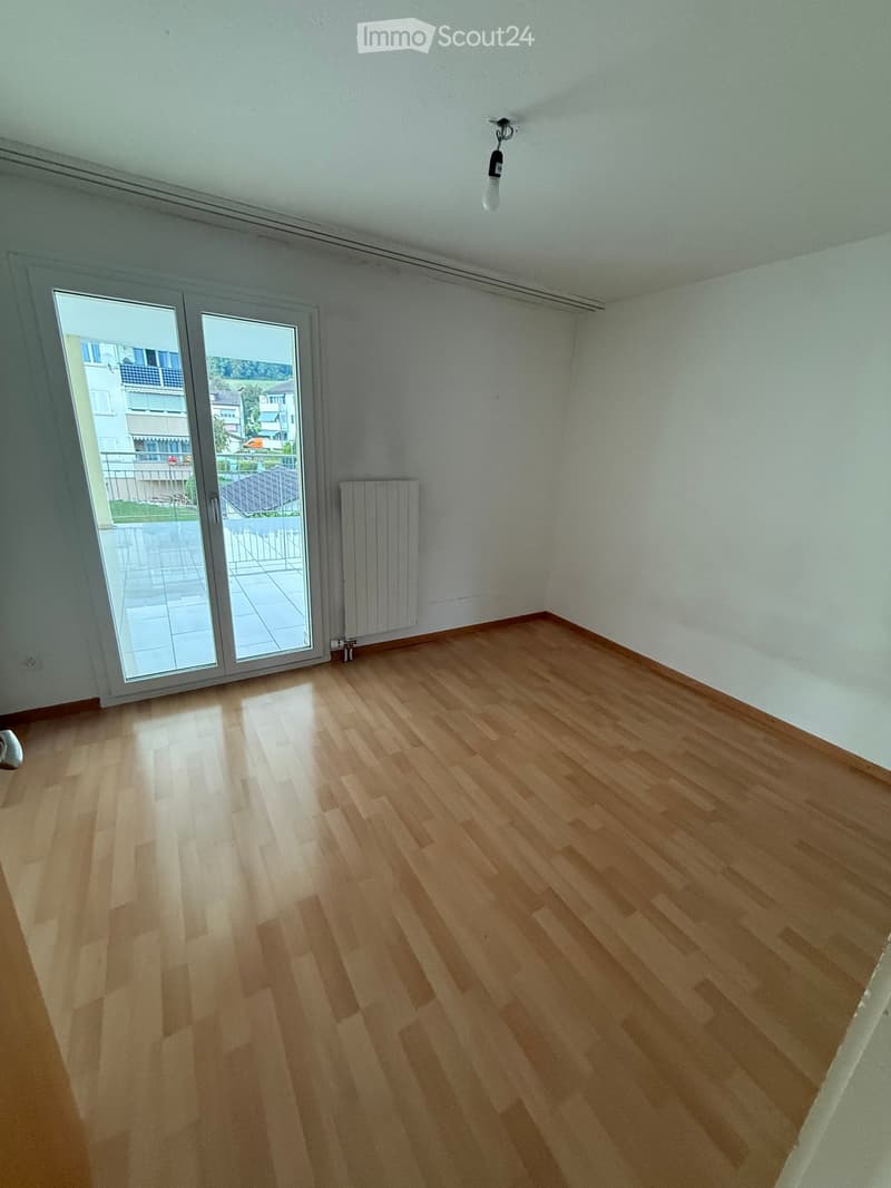 Empty room with wooden flooring, white walls, large sliding glass door leading to a balcony, radiator, and a single light fixture on the ceiling.