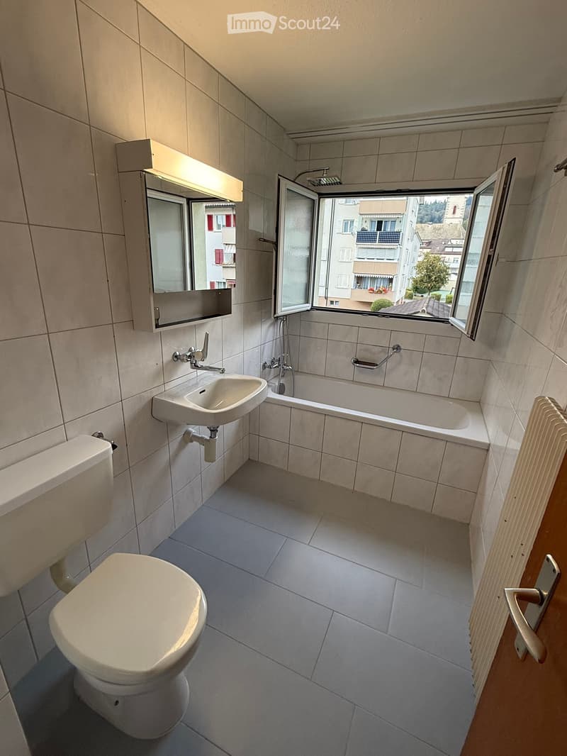 Modern bathroom with a toilet, sink, bathtub, mirror, and radiator. The room features tiled walls and floor, with an open window showing a view of the city.