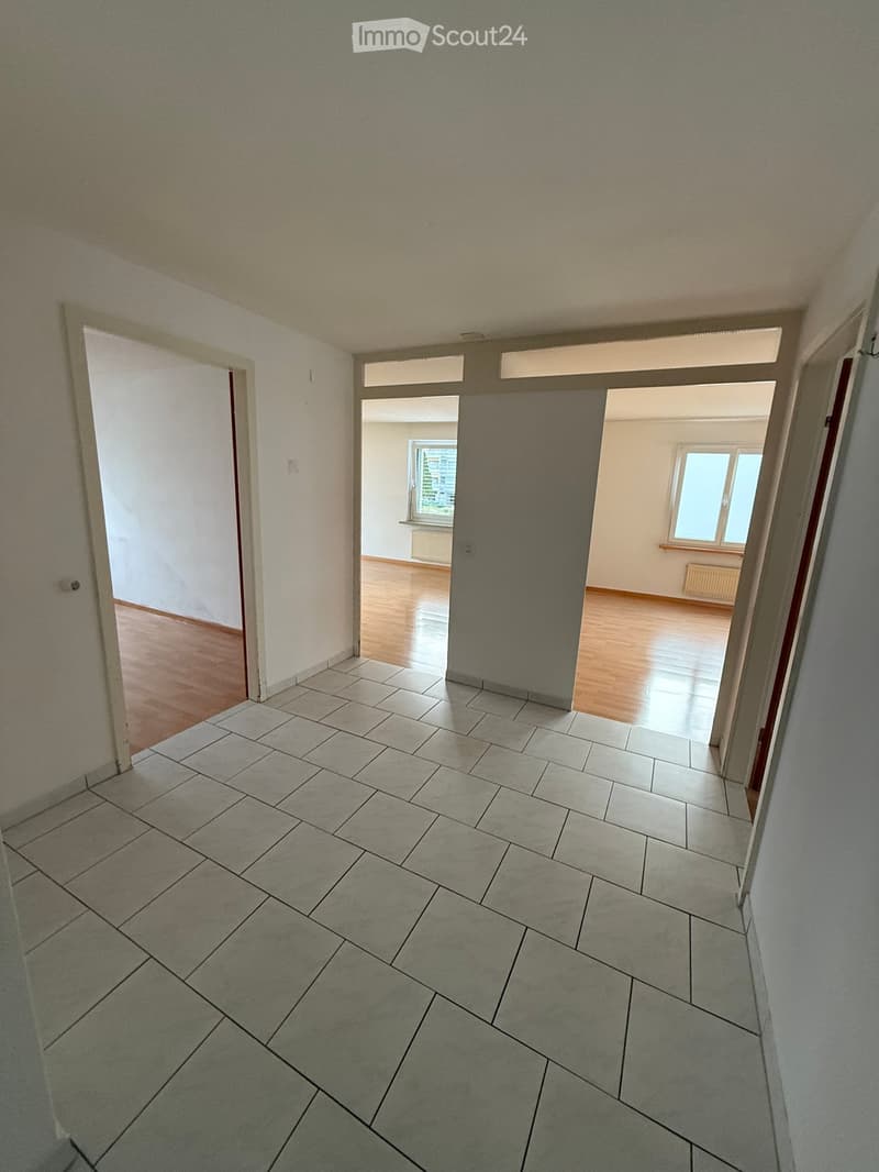 Empty hallway with tiled floor, wooden flooring in adjacent rooms, and white walls.