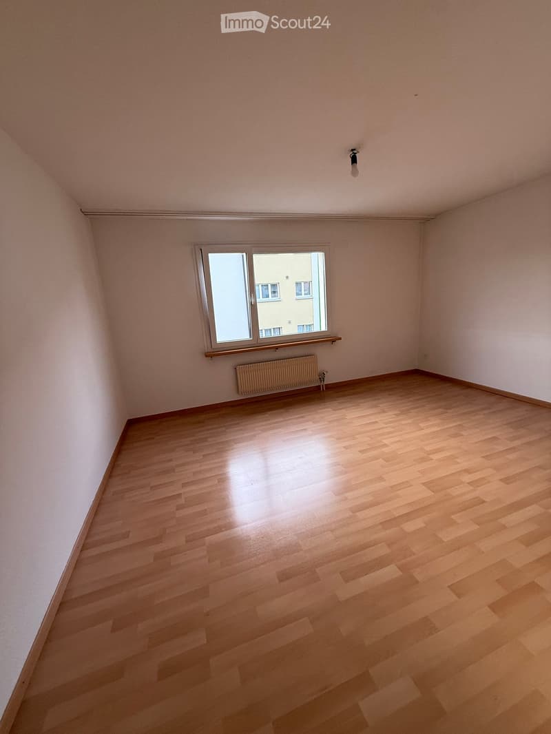 Empty room with light wooden flooring, white walls, and a single window with a radiator below it.