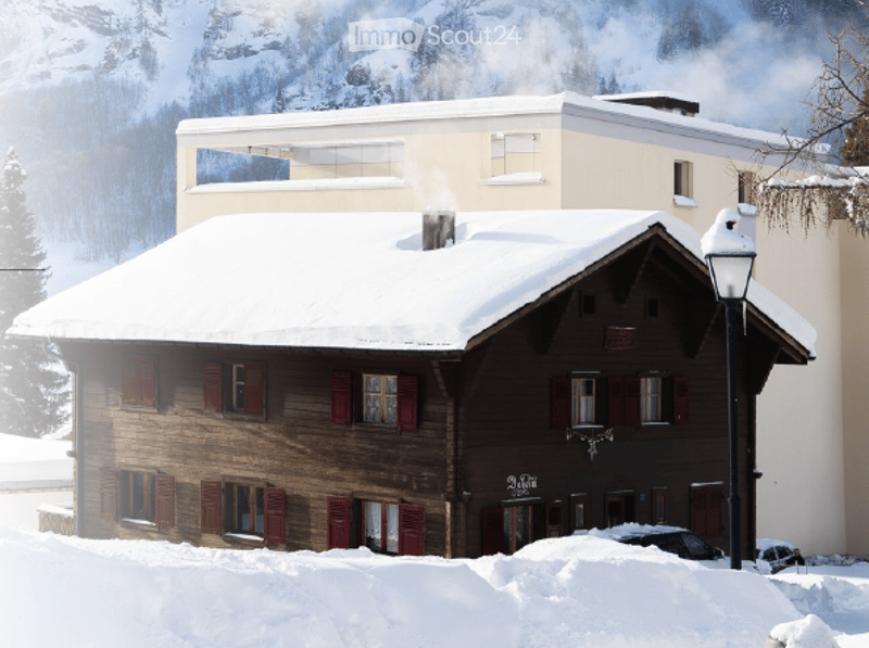 A traditional wooden chalet with red shutters and snow-covered roof, situated in a snowy mountain landscape with a modern building in the background.