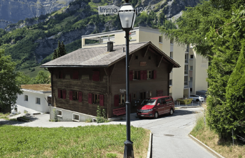 Traditional wooden chalet with red shutters and a grey roof, situated in a scenic mountainous area with a parked van nearby.
