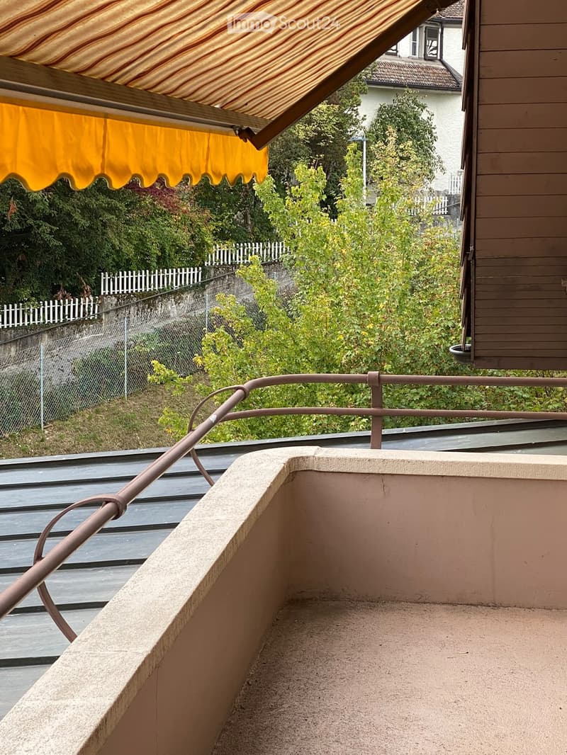 Balcony with metal railing, beige concrete floor, and a yellow striped awning extending from the building.