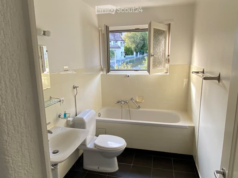 Bathroom with white tiles, a bathtub, toilet, sink, and a mirror. The window offers a view of the outside.
