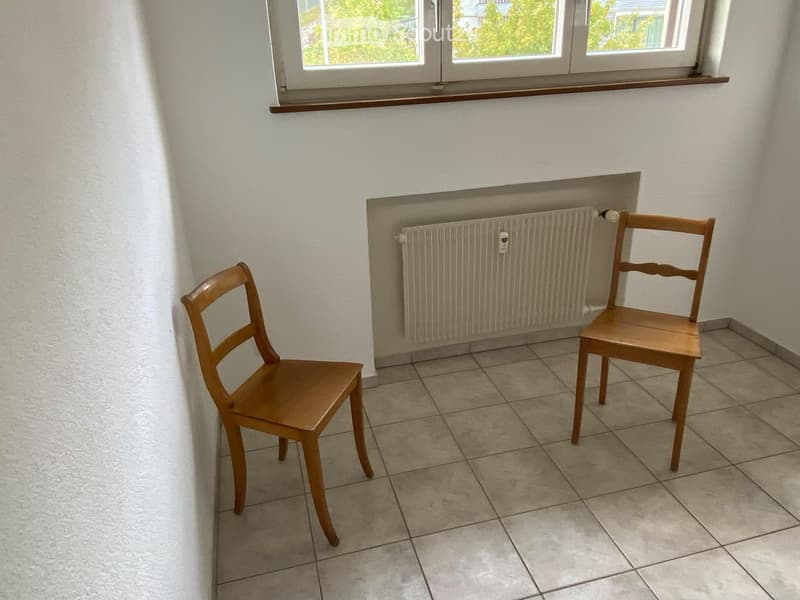 Empty room with tiled floor, two wooden chairs, radiator, and windows with wooden sills.