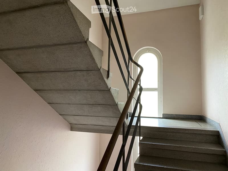 Staircase with concrete steps and a wooden handrail, leading to an arched window that allows natural light into the hallway.