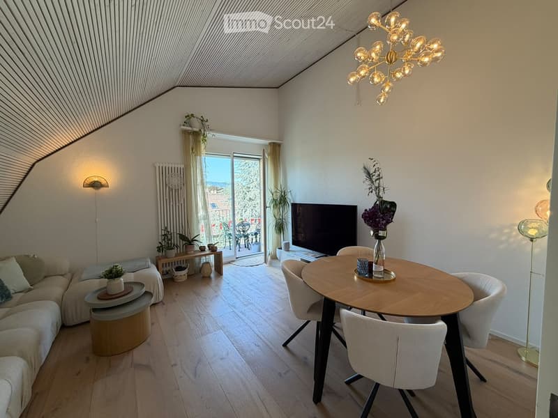Modern living and dining area with a slanted ceiling, large windows opening to a balcony, a sectional sofa, a coffee table, a dining table with chairs, a TV, and decorative plants.