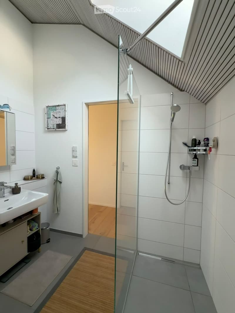 Modern bathroom with a glass-enclosed shower, white tiles, a sink with a mirror, and a towel hanging on the wall. The room features a sloped ceiling with skylights and a small wooden floor area visible through the open door.