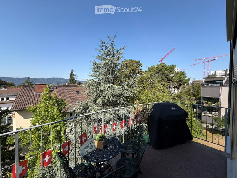 Balcony with metal railing, small round table, four chairs, potted plant, and covered grill. Swiss flags are attached to the railing. The view includes trees, rooftops, and distant mountains.
